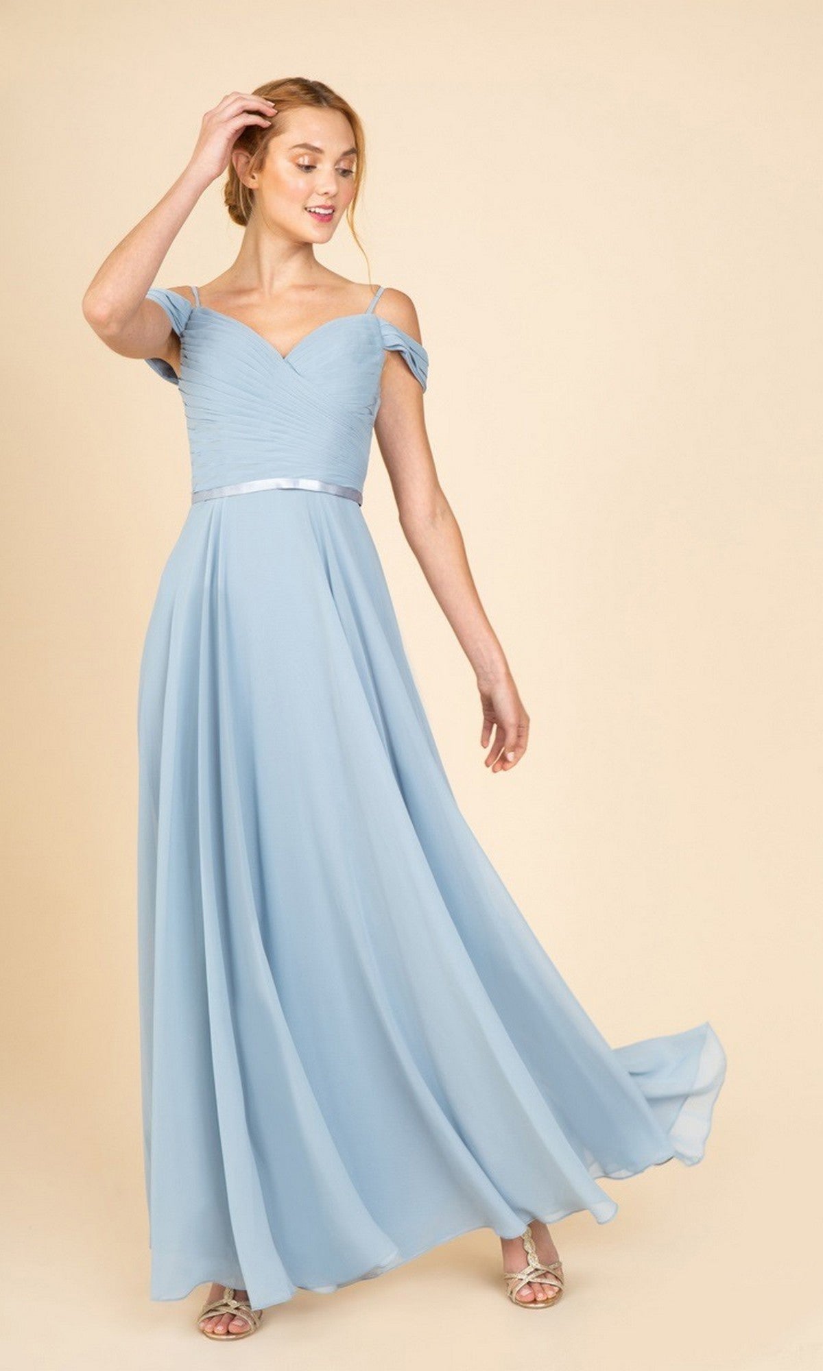 A woman stands against a beige background wearing the Eureka 7611 long prom dress in dusty blue, featuring an off-the-shoulder chiffon design with a ruched fitted bodice and flowing skirt.